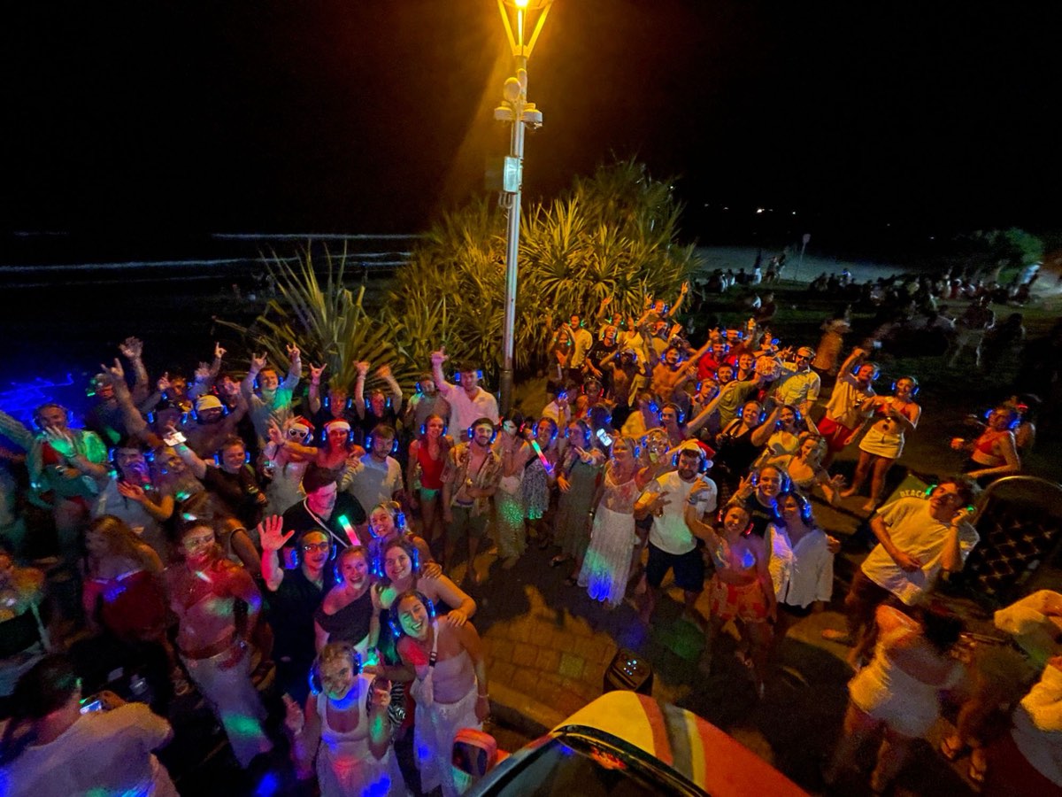 Big group in glowing headphones waving at the camera by the beach